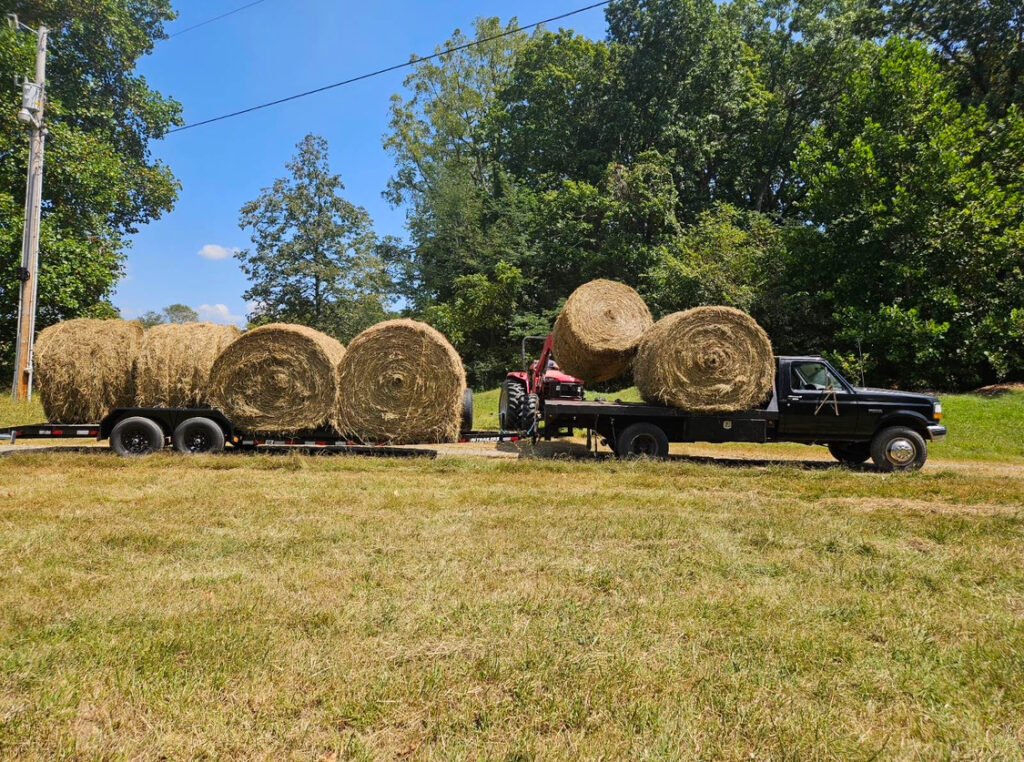 Large round hay bales on a trailer beside a red tractor in a freshly cut field at Retreat at Hope Farm in Springfield, Tennessee.