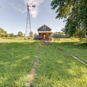 A rustic wooden cabin with a small porch sits in the middle of a sunlit grassy field, flanked by mature trees. A tall metal windmill stands beside the cabin, and faint tire tracks lead toward it under a bright blue sky with scattered clouds.