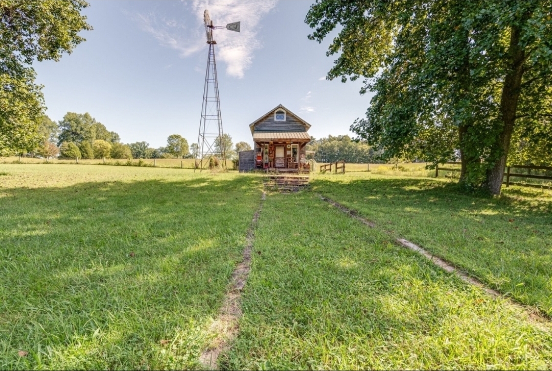A rustic wooden cabin with a small porch sits in the middle of a sunlit grassy field, flanked by mature trees. A tall metal windmill stands beside the cabin, and faint tire tracks lead toward it under a bright blue sky with scattered clouds.