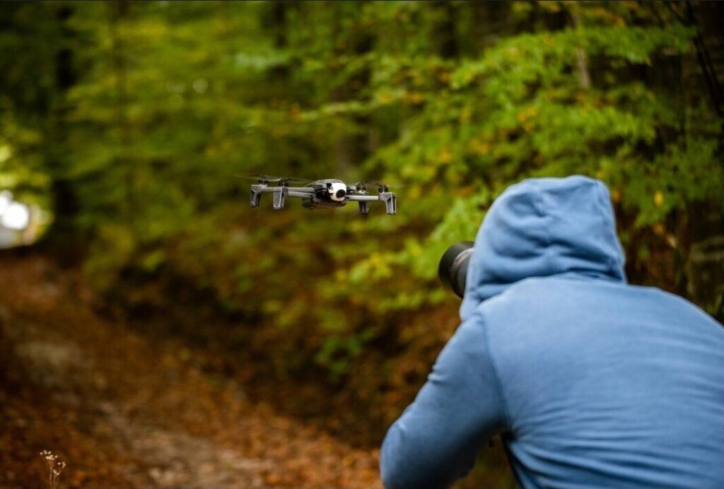 A person in a blue hoodie photographing a drone flying through a forest trail with green and autumn foliage