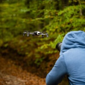 A person in a blue hoodie photographing a drone flying through a forest trail with green and autumn foliage