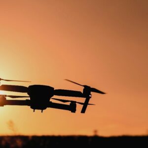 A drone flying in silhouette against an orange sunset sky with the sun low on the horizon.