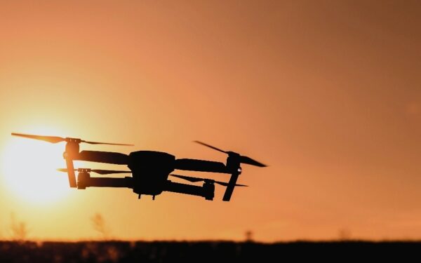 A drone flying in silhouette against an orange sunset sky with the sun low on the horizon.