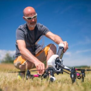 A man wearing sunglasses kneels in a field while preparing a large quadcopter drone for flight on a sunny day.
