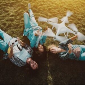 A group of friends lying on the grass laughing as a drone hovers above them during a sunny outdoor flying session.