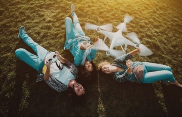 A group of friends lying on the grass laughing as a drone hovers above them during a sunny outdoor flying session.